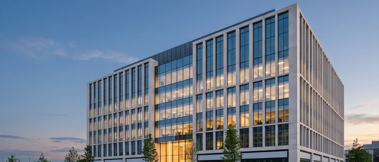 A seven-storey Class A office building at blue hour: pale limestone fins, a floor-to-ceiling glass curtain wall with warm-lit floors, a glowing double-height lobby and a wide stone plaza in front.